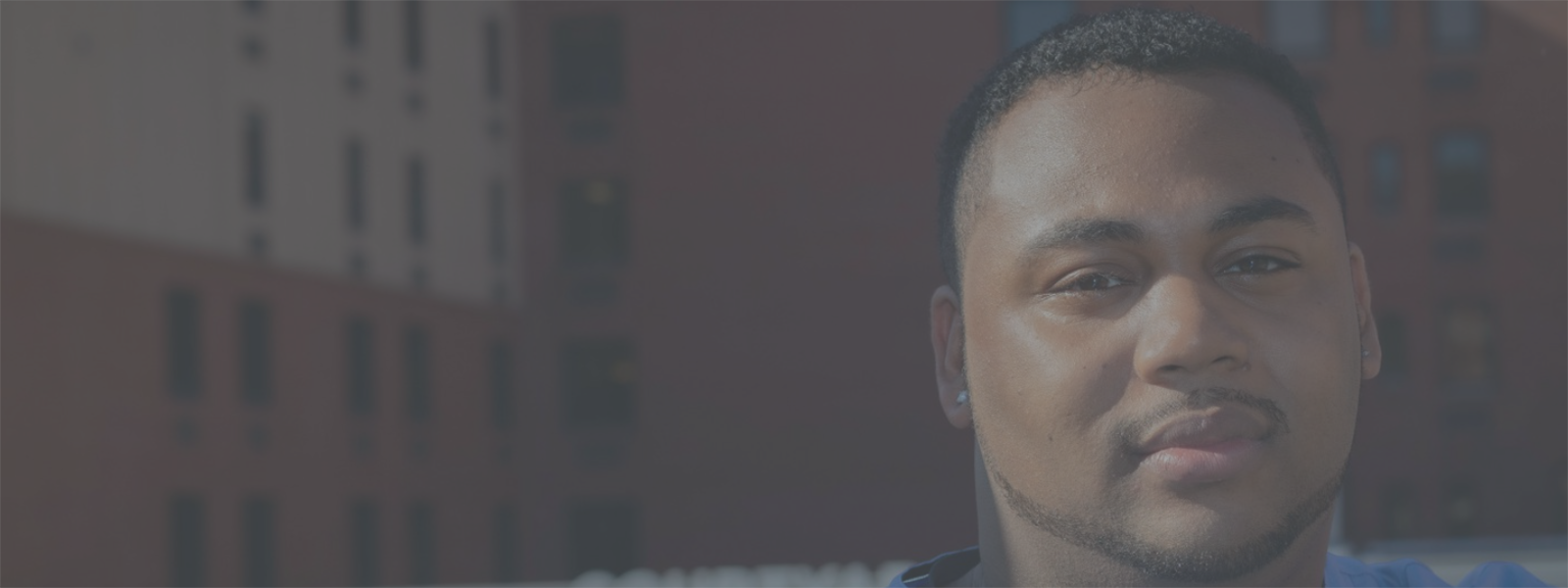 Photo of a Black male nurse in blue scrubs, facing the camera and smiling slightly