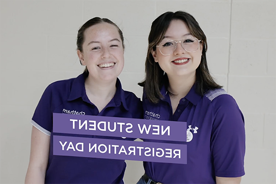 Two student volunteers wearing purple smile together for a photo. 'New Student Registration Day' is written on a purple banner in front of them.