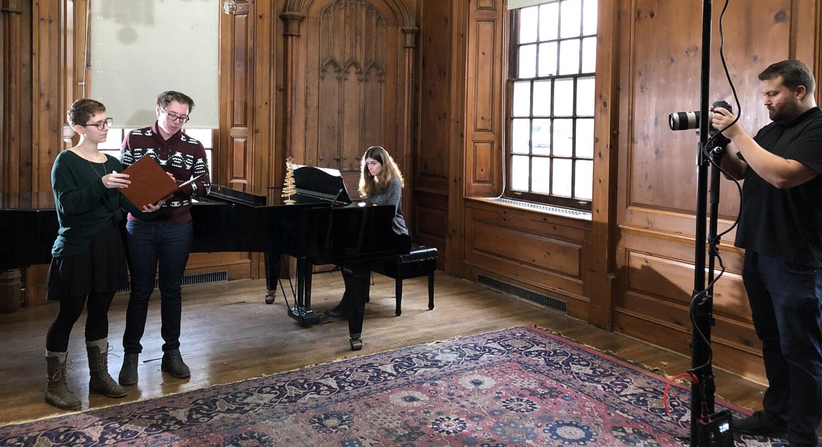 Photo of two students standing and singing, with a woman playing a grand piano behind them. They are being filmed by a person on the left of the photo.